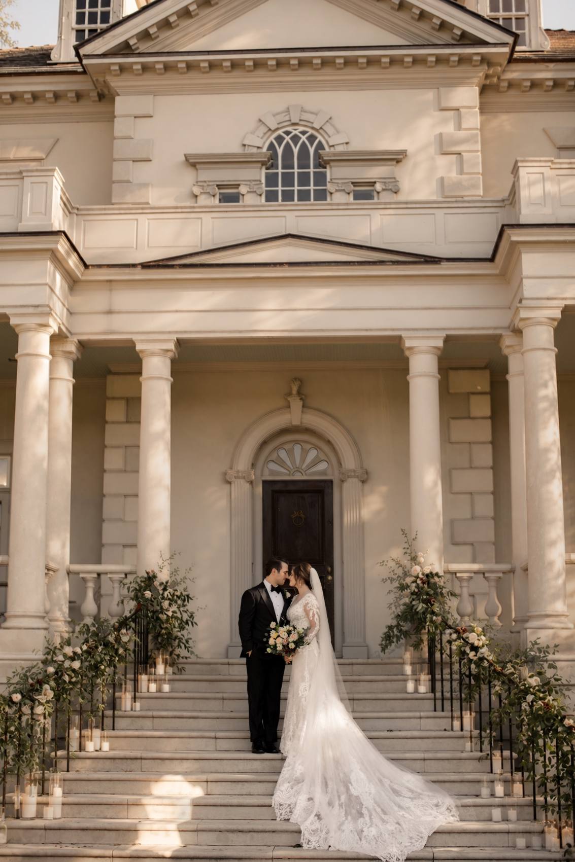 Bride in a long lace gown and veil and groom in a black tuxedo standing forehead to forehead on the front steps of a columned mansion, flower garlands and candles along the iron railings