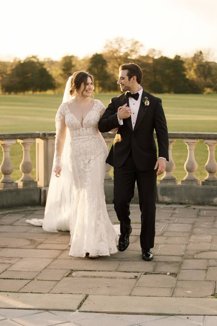Bride and groom walking arm in arm across a stone terrace in front of a balustrade and open lawn at golden hour
