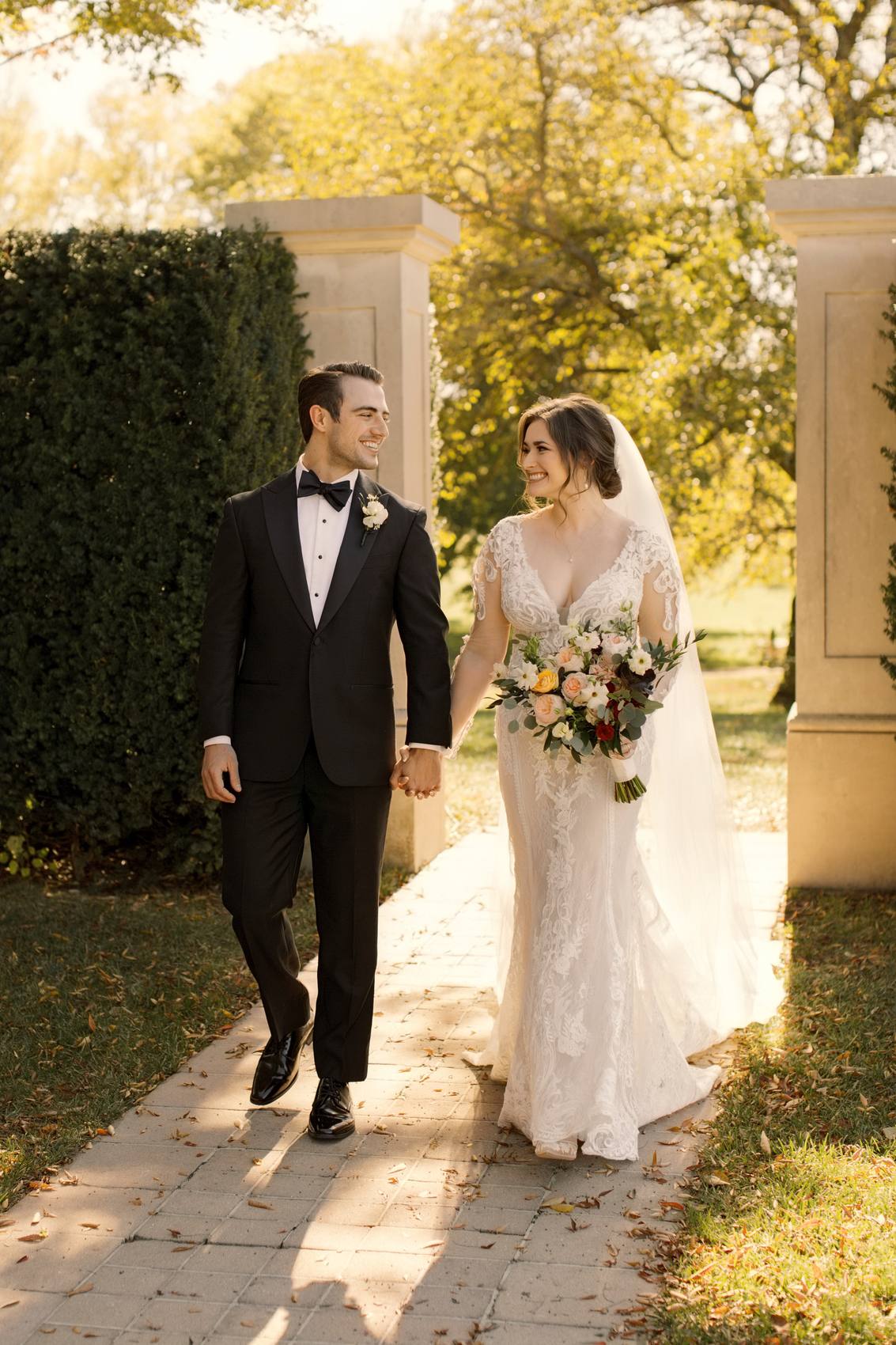 Bride and groom walking hand in hand toward the camera on a brick path between stone pillars and a clipped hedge, golden autumn trees behind them