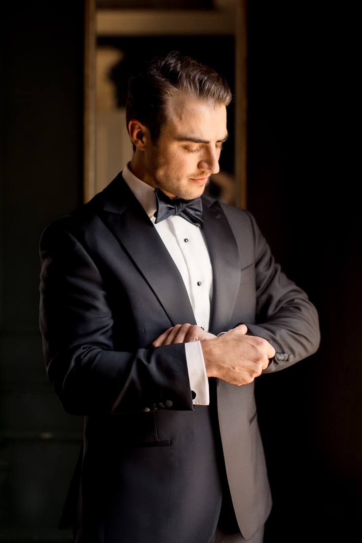 Groom in a black tuxedo looking down as he adjusts his cuff in dim window light