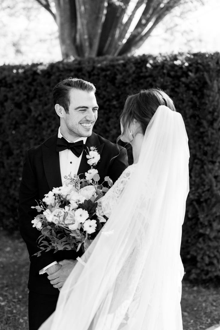 Black and white close photo of the groom smiling at the bride, her veil in the foreground, a clipped hedge behind them