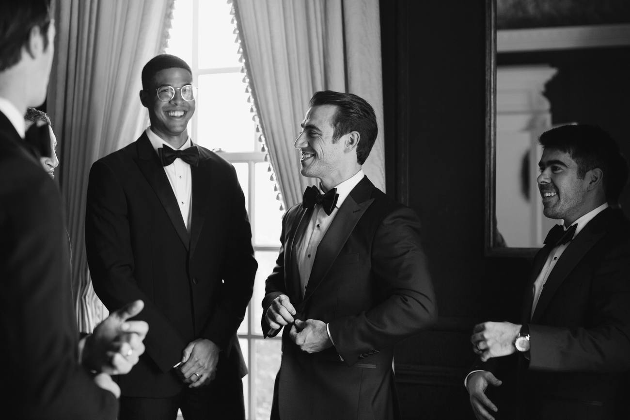 Black and white photo of groomsmen in tuxedos laughing together beside a tall window with long curtains