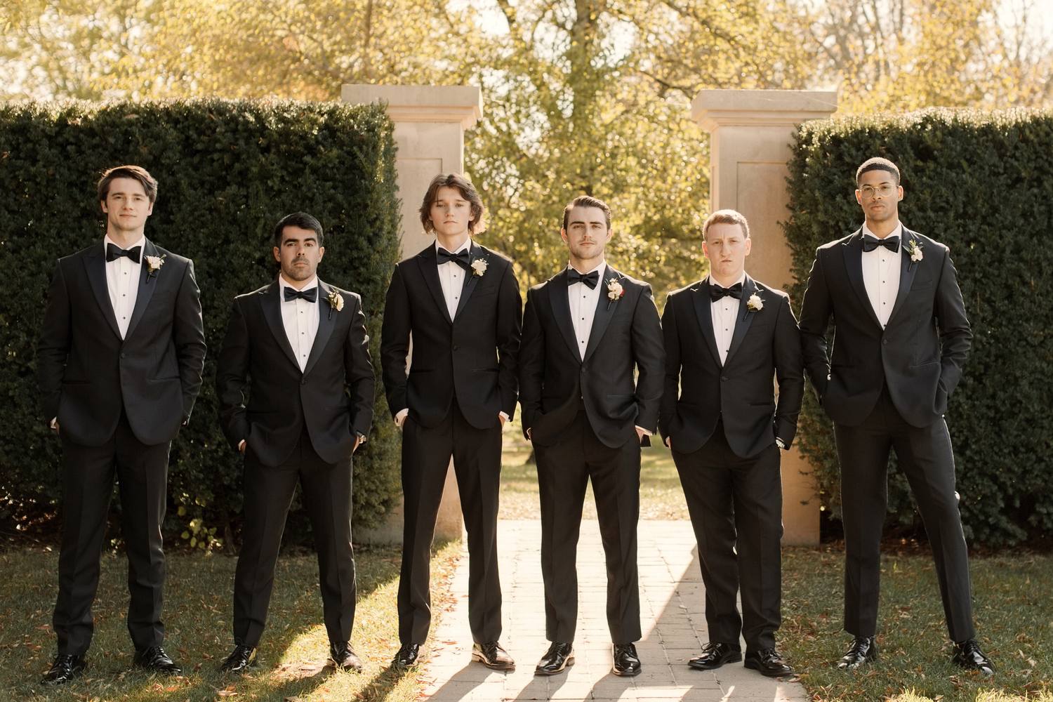 Six groomsmen in black tuxedos and bow ties standing in a line on a brick path between stone pillars and tall clipped hedges