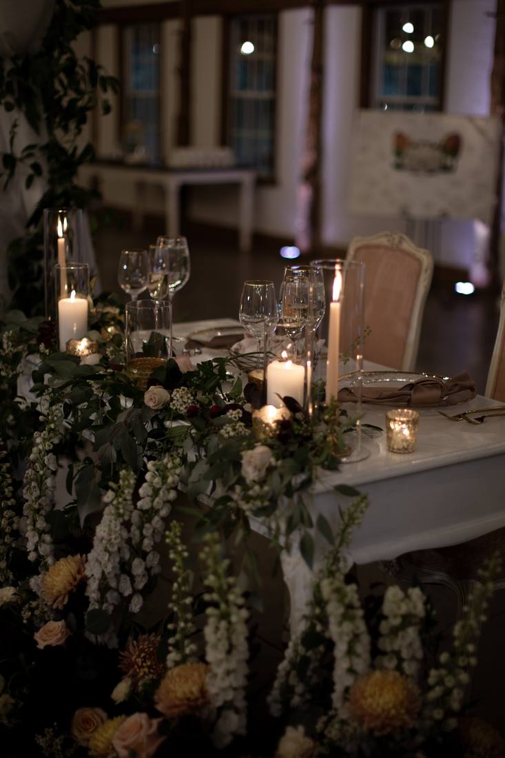 Sweetheart table with a tumble of white delphinium, roses and dahlias, tall candles, glassware and blush upholstered chairs in a timber-framed reception room