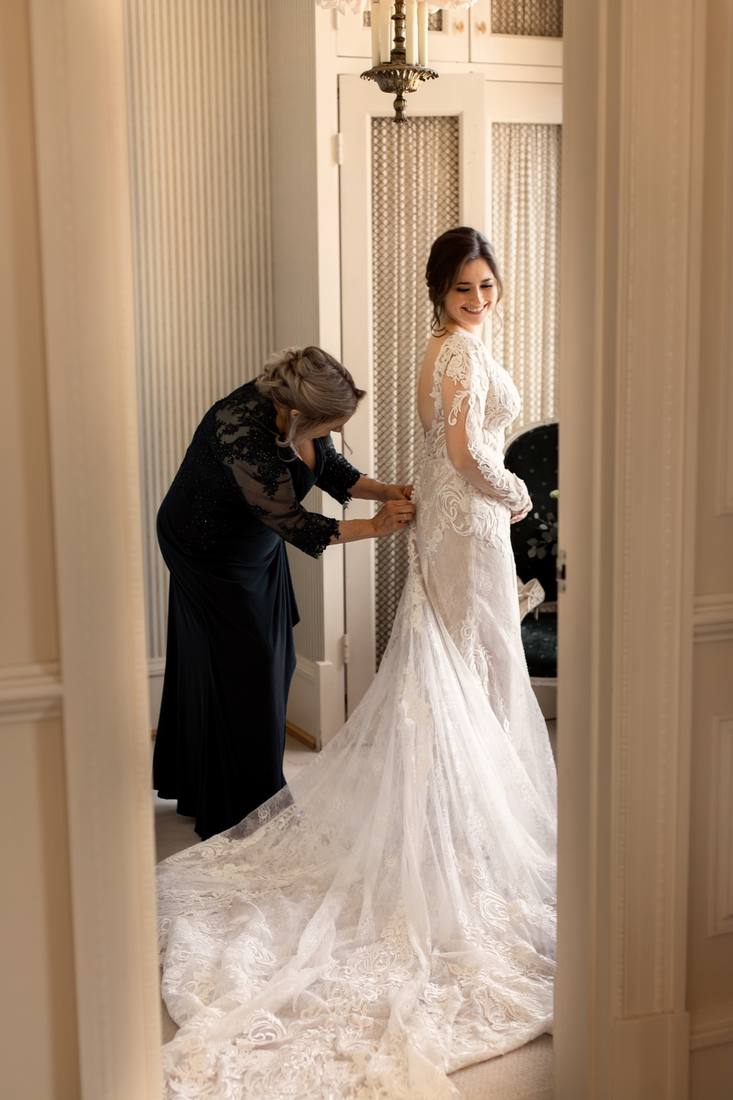 A woman in black helping the bride with the back of her lace gown, the long train spread across the floor of a tall room