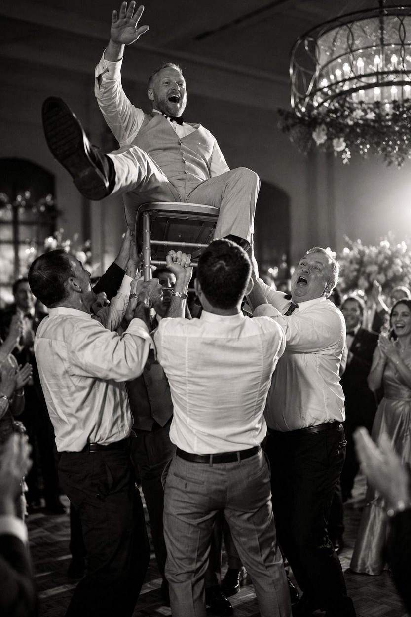 Black-and-white photograph of a groom raised on a chair above the dance floor, arm in the air, as guests lift him and cheer.
