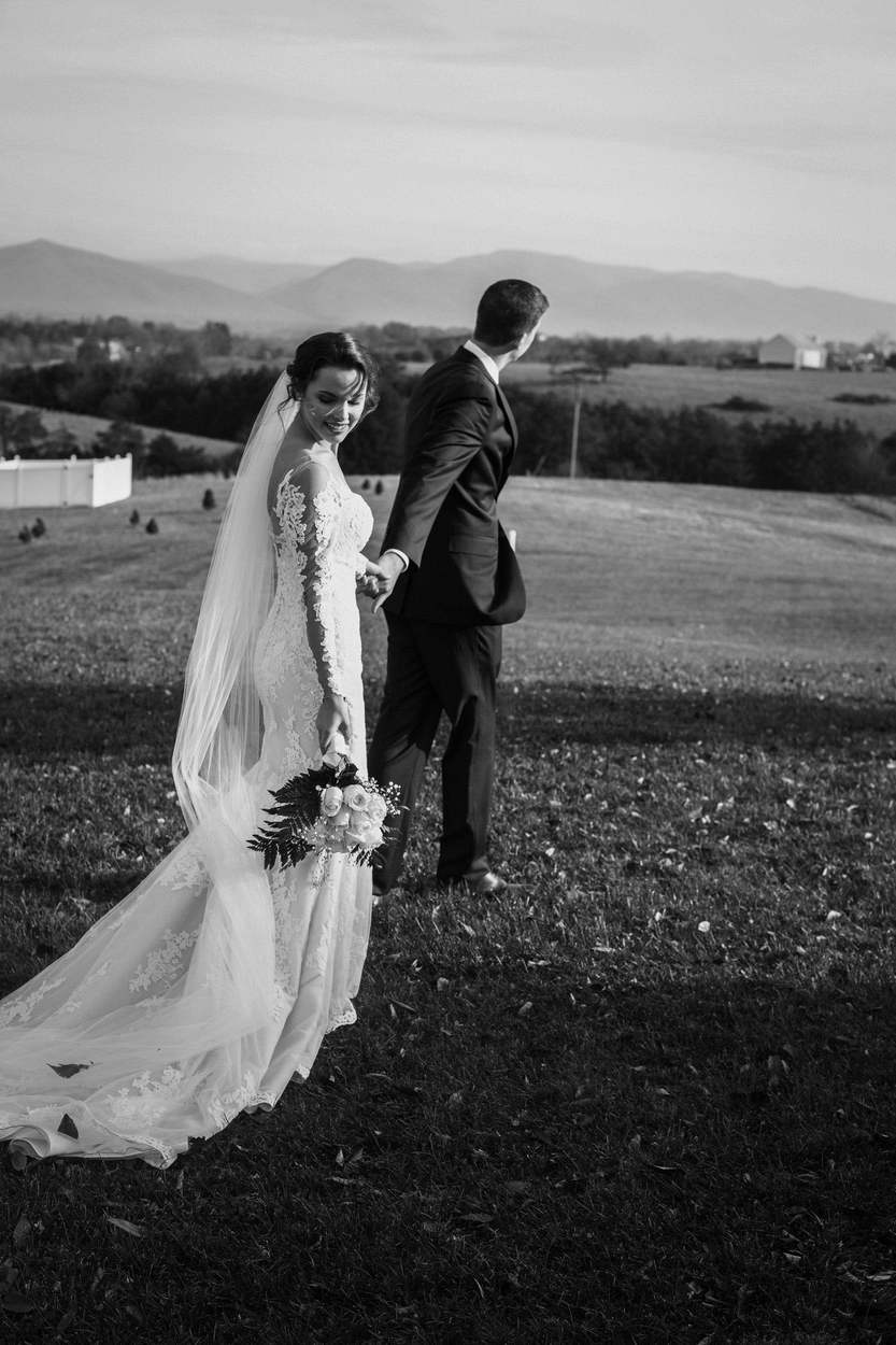 Black-and-white photograph of a bride glancing down with a smile as she holds her groom's hand and a bouquet of roses, walking across a hillside with distant mountains.