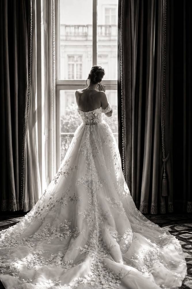 Bride in a lace gown seen from behind at a tall window between heavy curtains, her long train spread across the floor