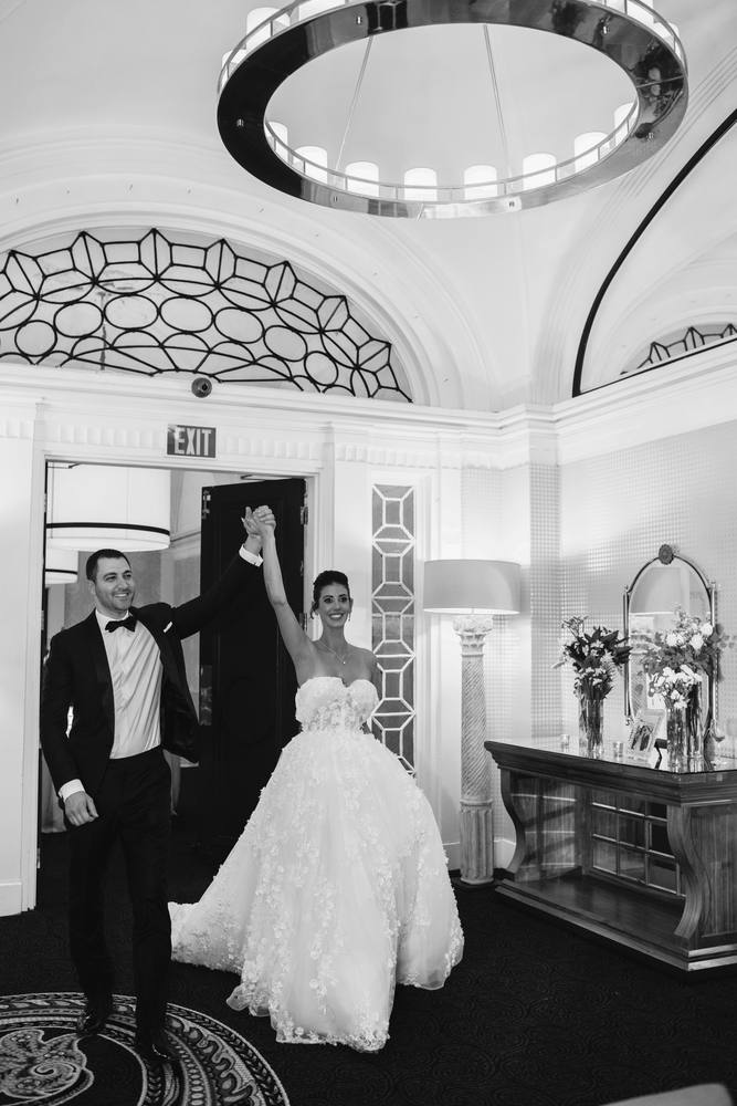 Black and white photo of a bride raising her fist as she enters a ballroom, men in tuxedos beside her under a chandelier