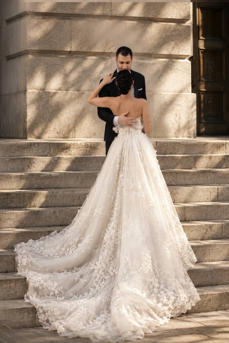 Bride seen from behind in a long lace gown with a flowing train, embracing the groom on sunlit stone steps