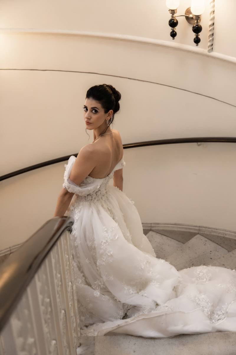 Bride in an off-the-shoulder lace gown glancing over her shoulder on a curved marble staircase with a dark wood handrail