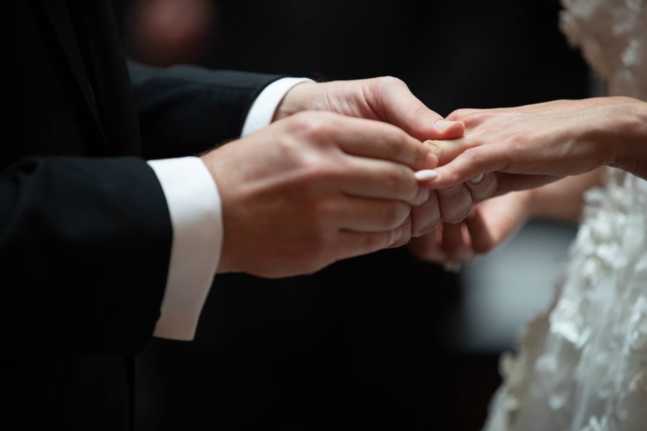 Close view of a ring exchange, his white cuff and black sleeve beside her hand and lace sleeve
