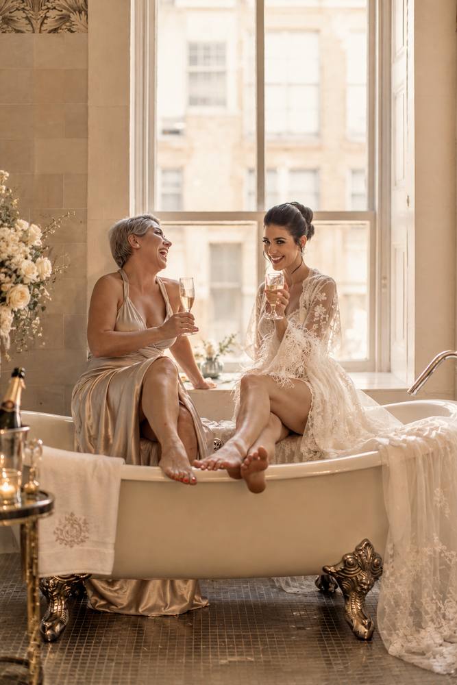Two women in robes laughing together in a clawfoot bathtub in a sunlit room with tall windows