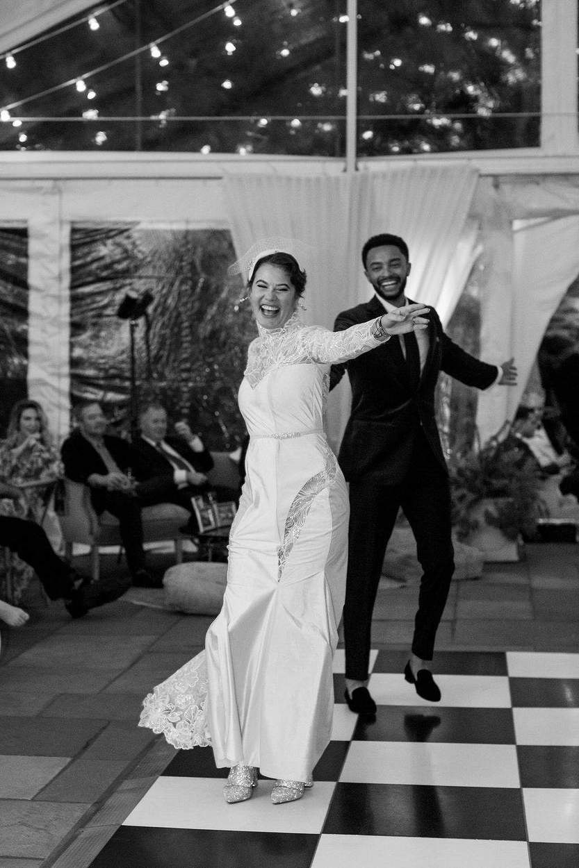 Black-and-white photograph of a laughing bride and groom stepping onto a checkered dance floor under string lights in a clear-sided tent, guests watching from sofas.