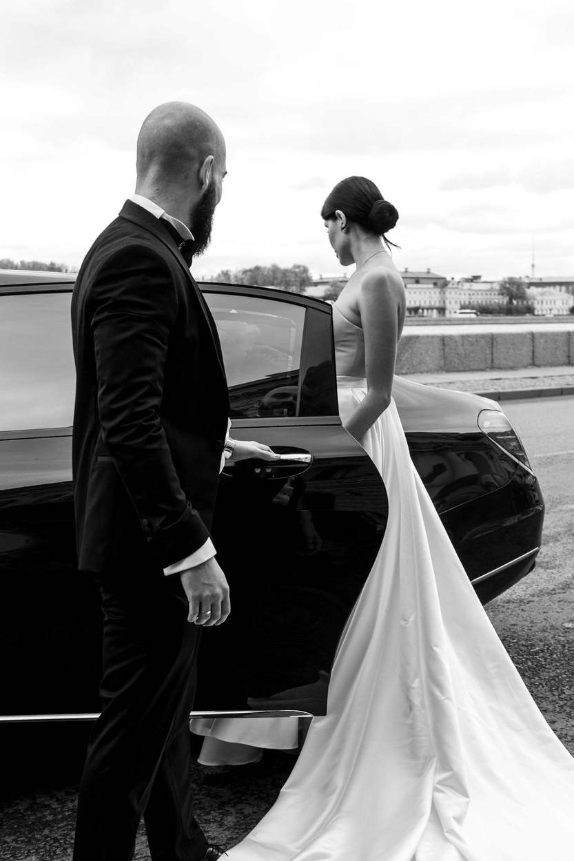Black-and-white photograph of a groom in a tuxedo holding open the door of a black car as his bride, in a long satin gown, steps in beside a river embankment.