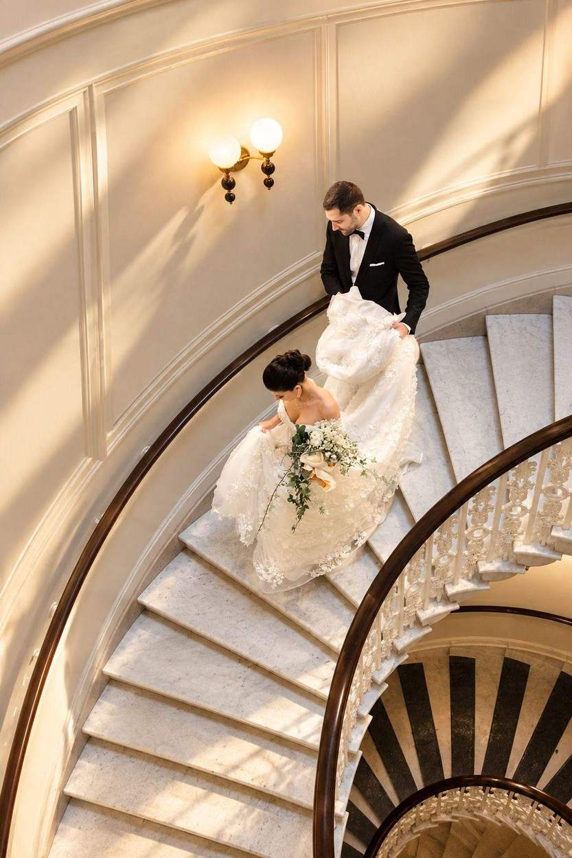 A groom in a black tuxedo carries the train of his bride's lace gown as they walk down a curved marble staircase, photographed from above.