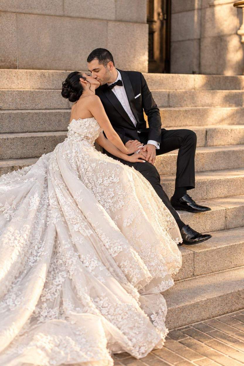 A bride seated on stone steps in a voluminous lace gown kisses her groom, his tuxedo and polished shoes framing the cascading train.