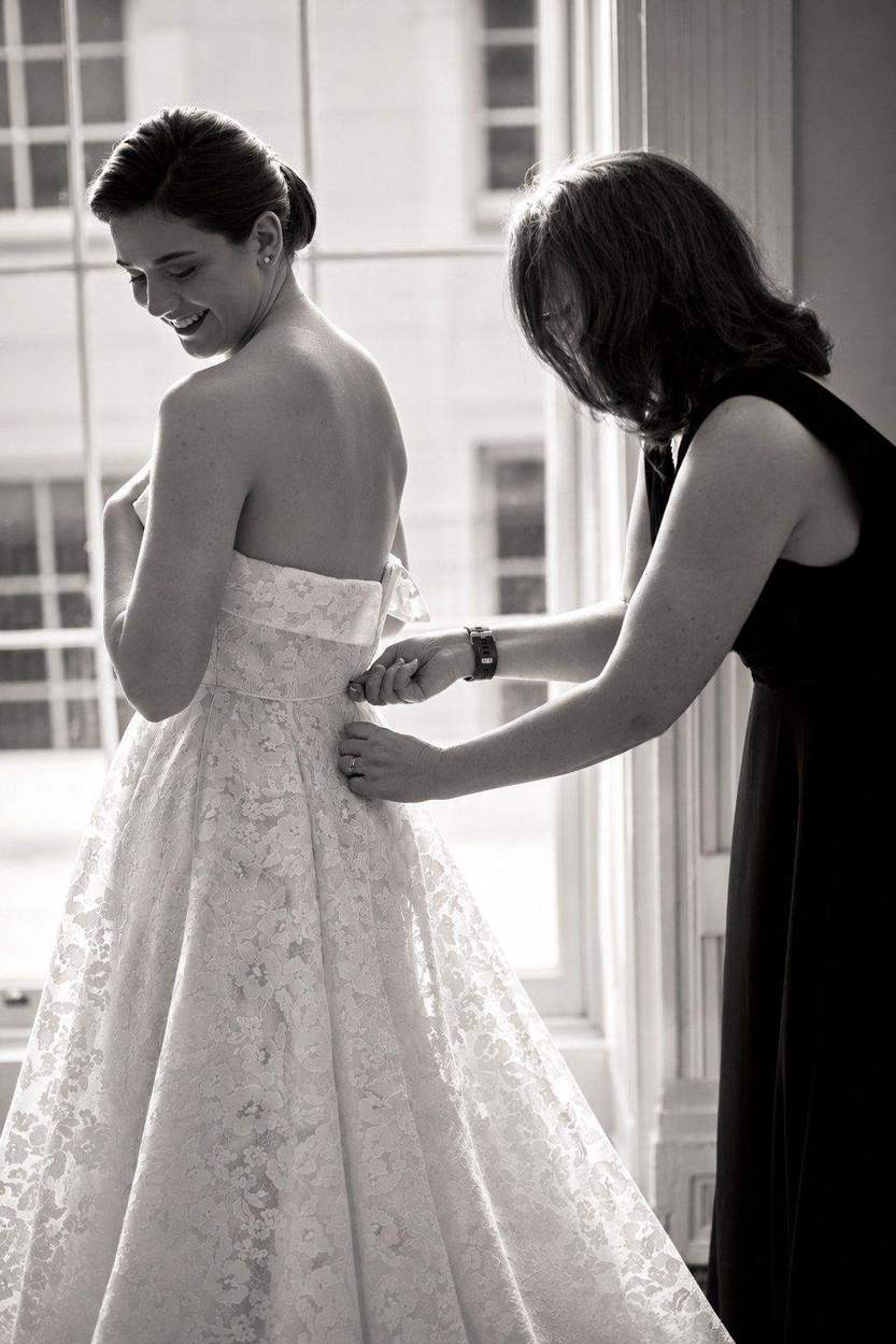 Washington DC photographer black and white bridal portrait: a smiling bride having her lace gown fastened by a bridesmaid beside a sunlit window.