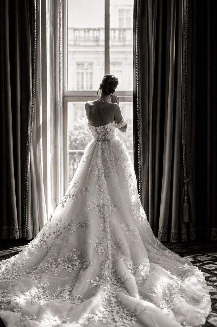 Washington DC photographer black and white bridal portrait: a bride seen from behind in a floral gown with a long train, standing at a tall window between velvet curtains.