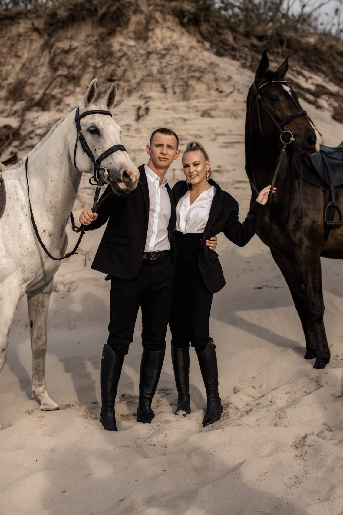 Smiling couple in black standing arm in arm between a white horse and a dark horse on a sand dune