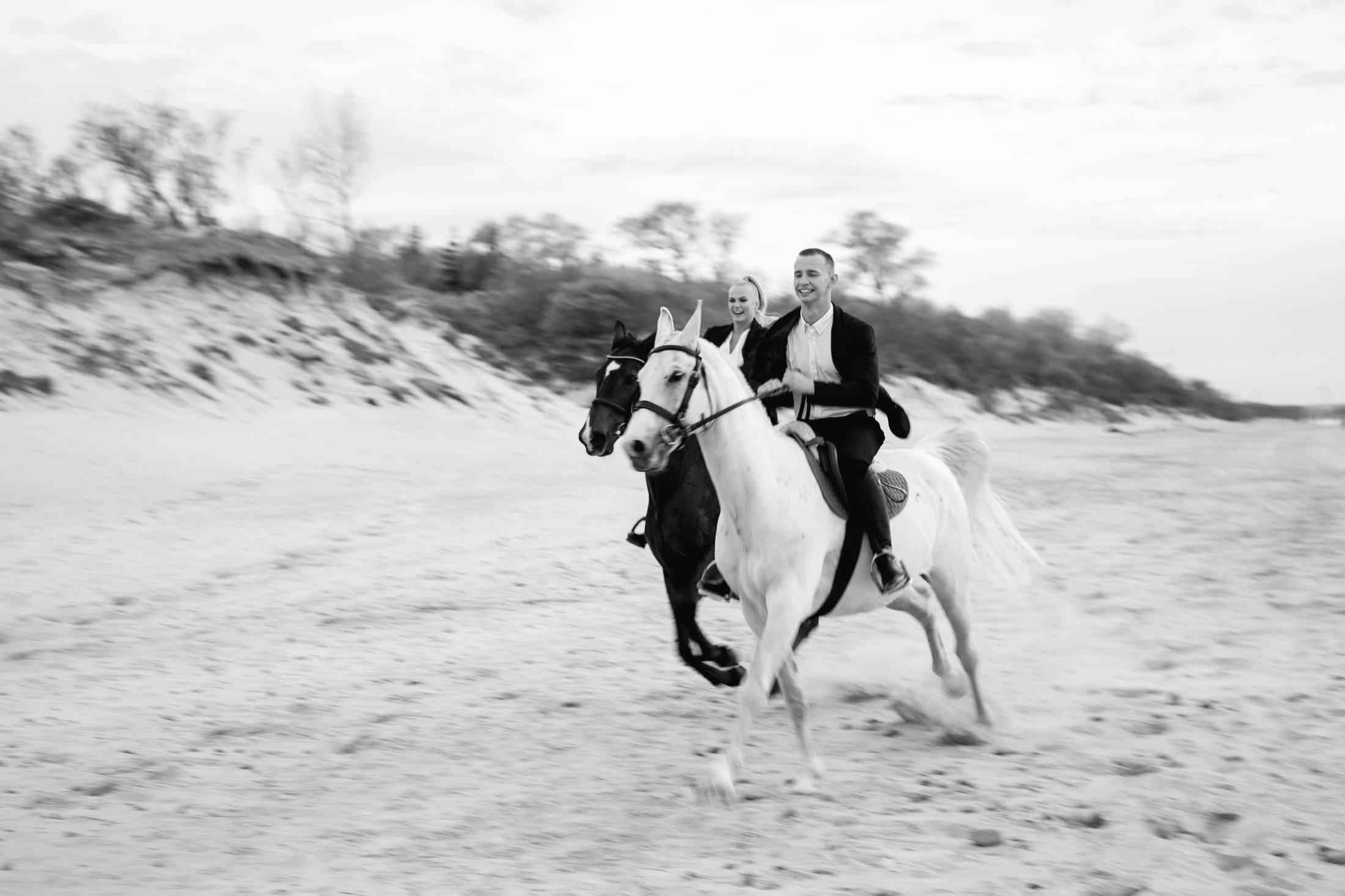 Black and white photograph of a couple galloping on a white horse across the sand, dunes and trees behind