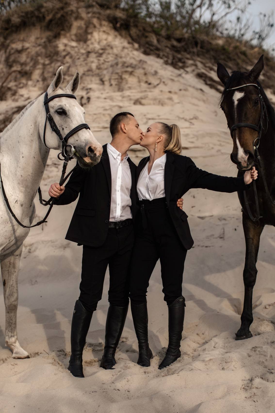 Couple in black kissing between a white horse and a dark horse on a sand dune