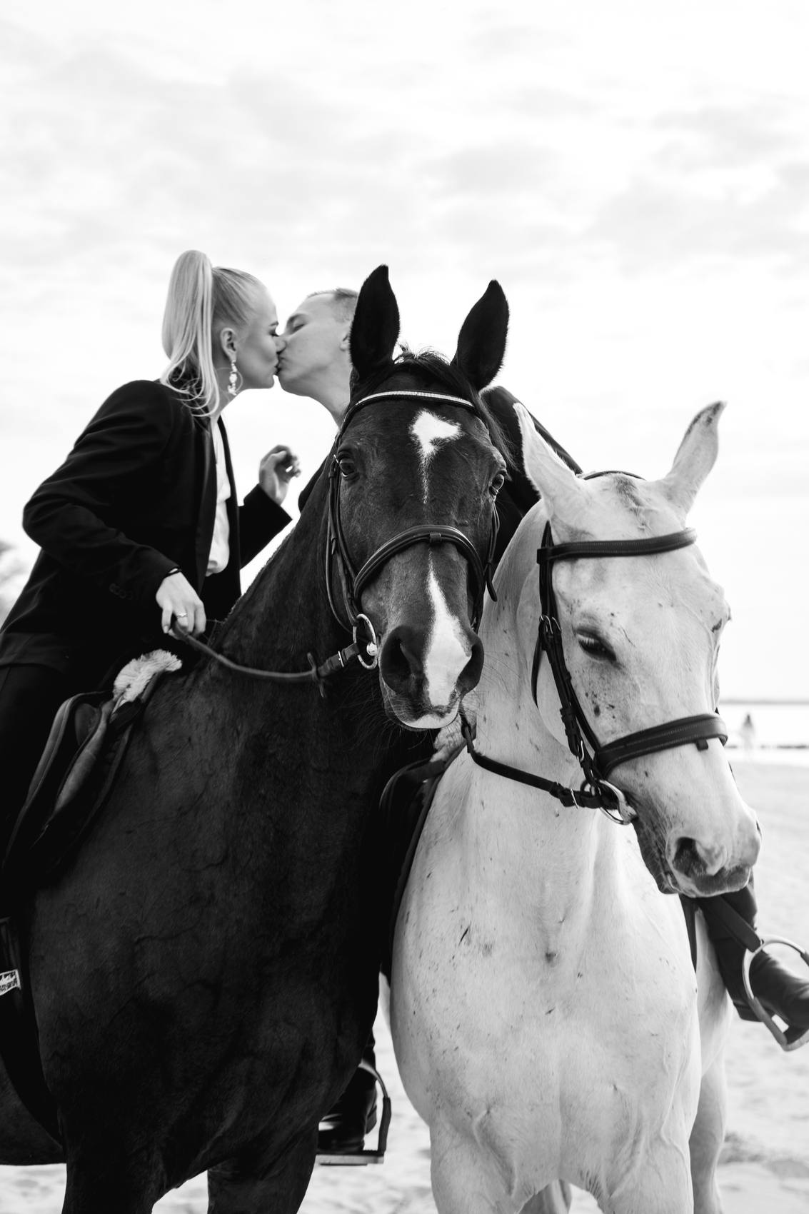 Black and white photograph of a couple kissing between a dark horse and a white horse