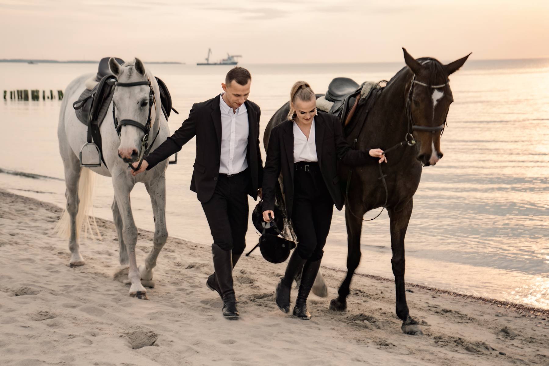 Couple in black leading a white horse and a dark horse along the shoreline in warm sunset light, a ship on the horizon