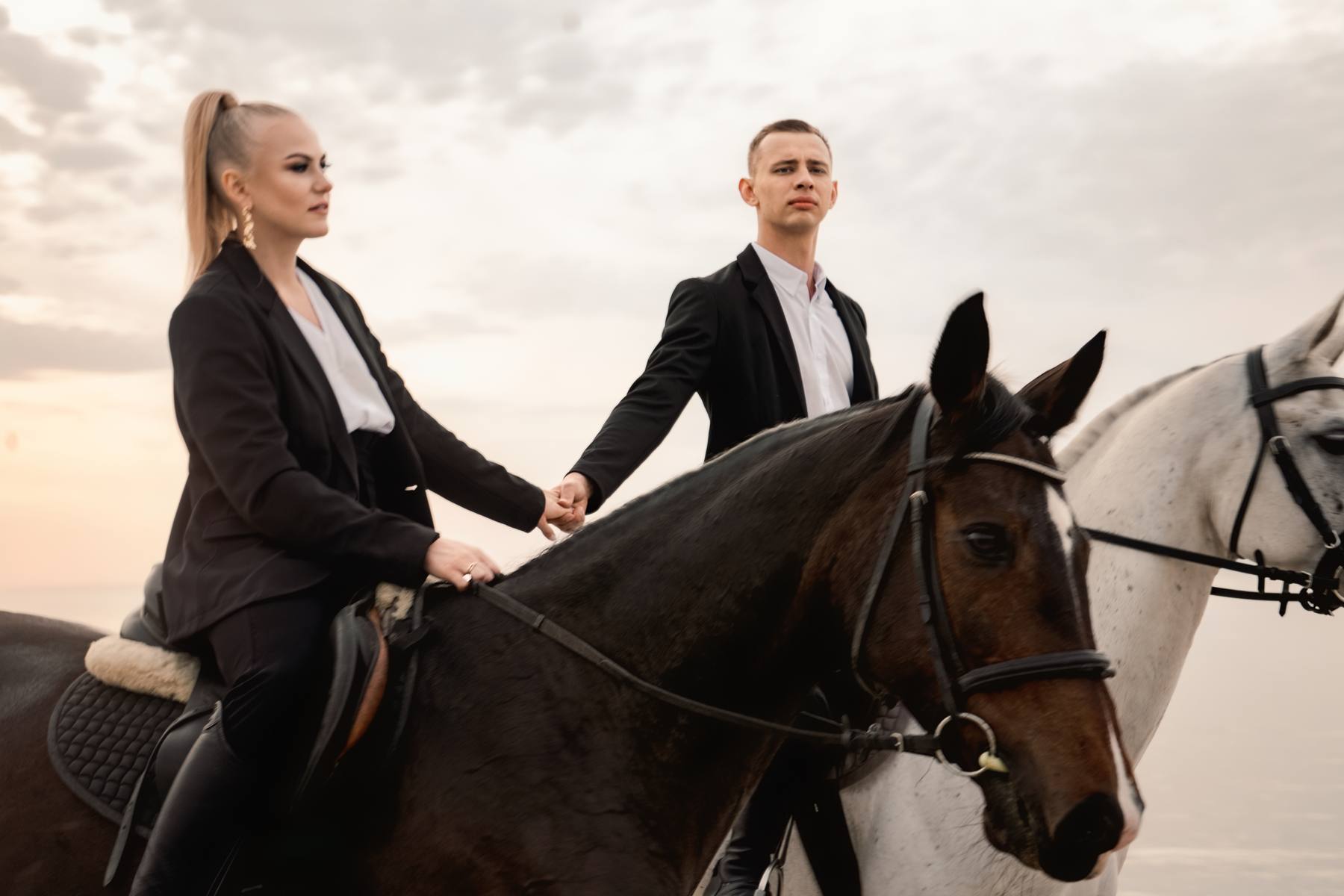 Woman and man in black blazers sitting on a dark bay horse holding hands, a white horse beside them, soft golden light in a pale sky