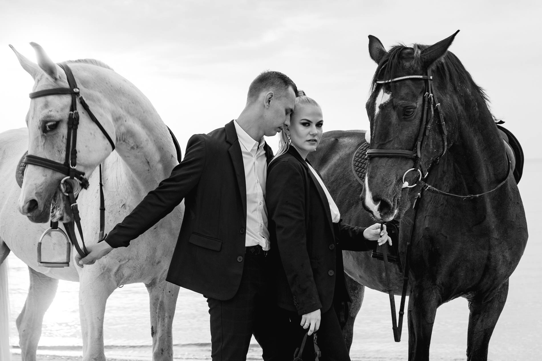 Black and white portrait of a couple in black standing between a white horse and a dark horse, the man reaching toward the white horse's bridle