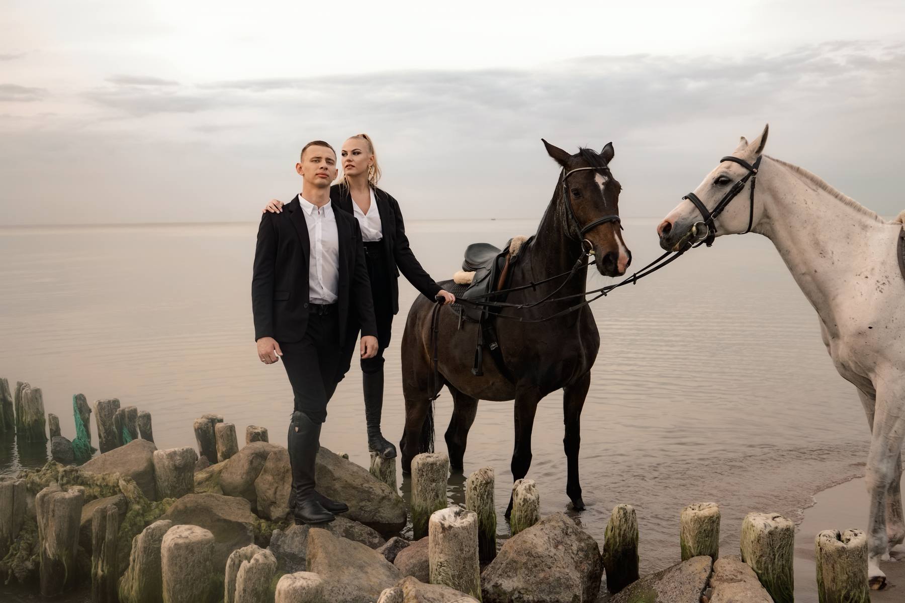 Couple in black standing on a stone and timber breakwater beside a dark bay horse and a white horse, calm water behind