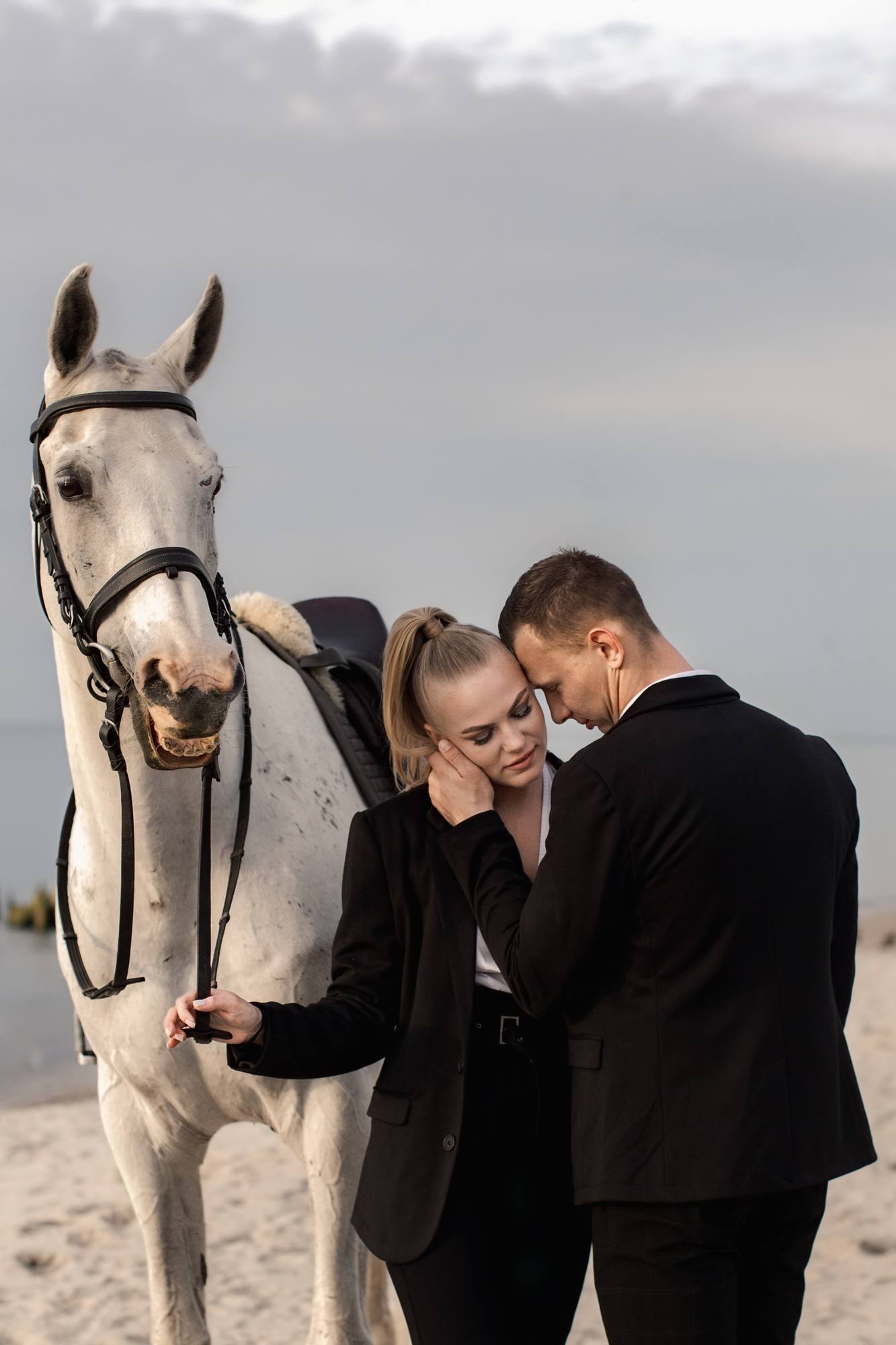 Engaged couple in black standing forehead to forehead on the beach while a smiling white horse leans in beside them, soft gray sky and water behind