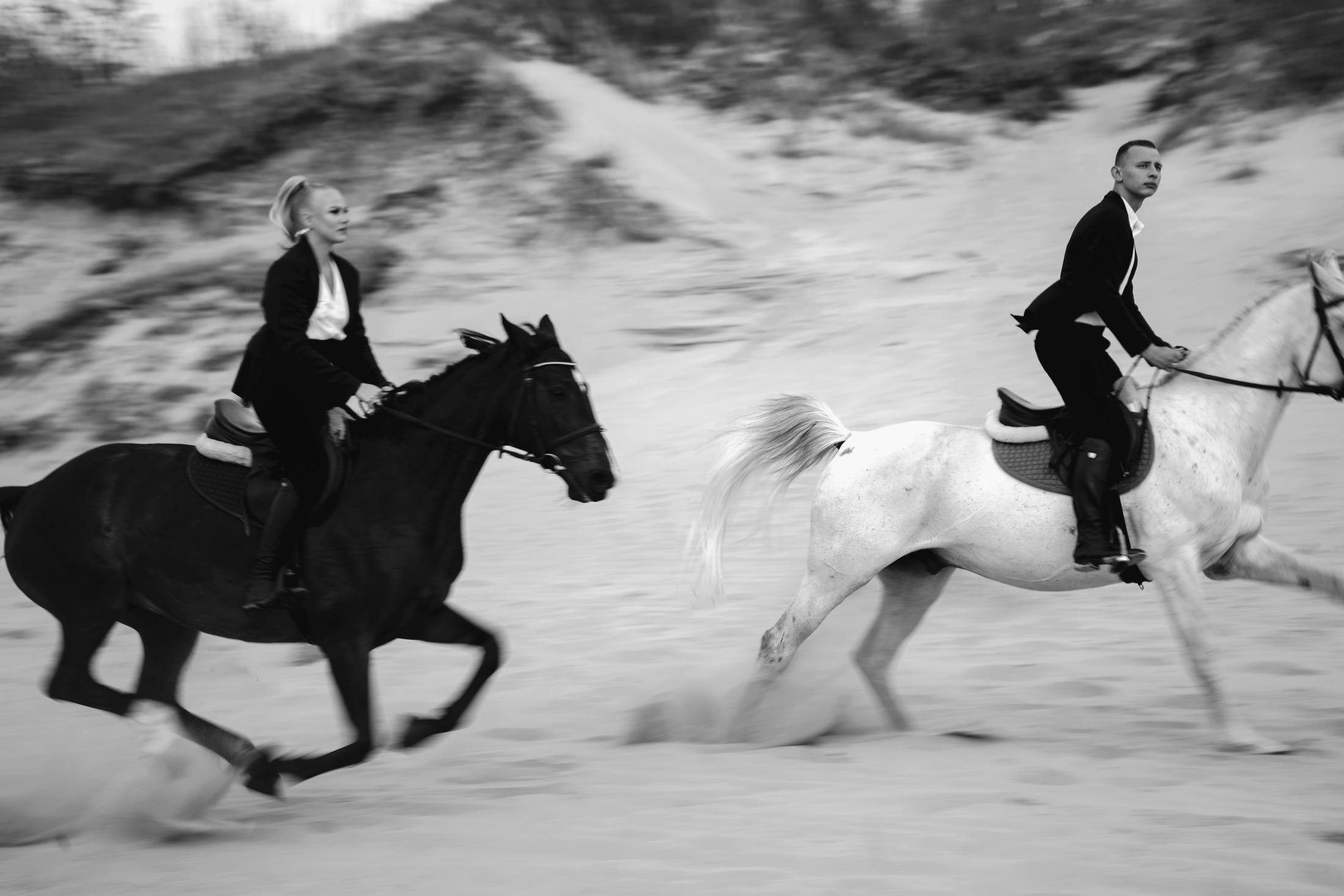 Black and white photograph of two riders galloping their horses across the sand, one dark horse and one white, dunes blurred behind