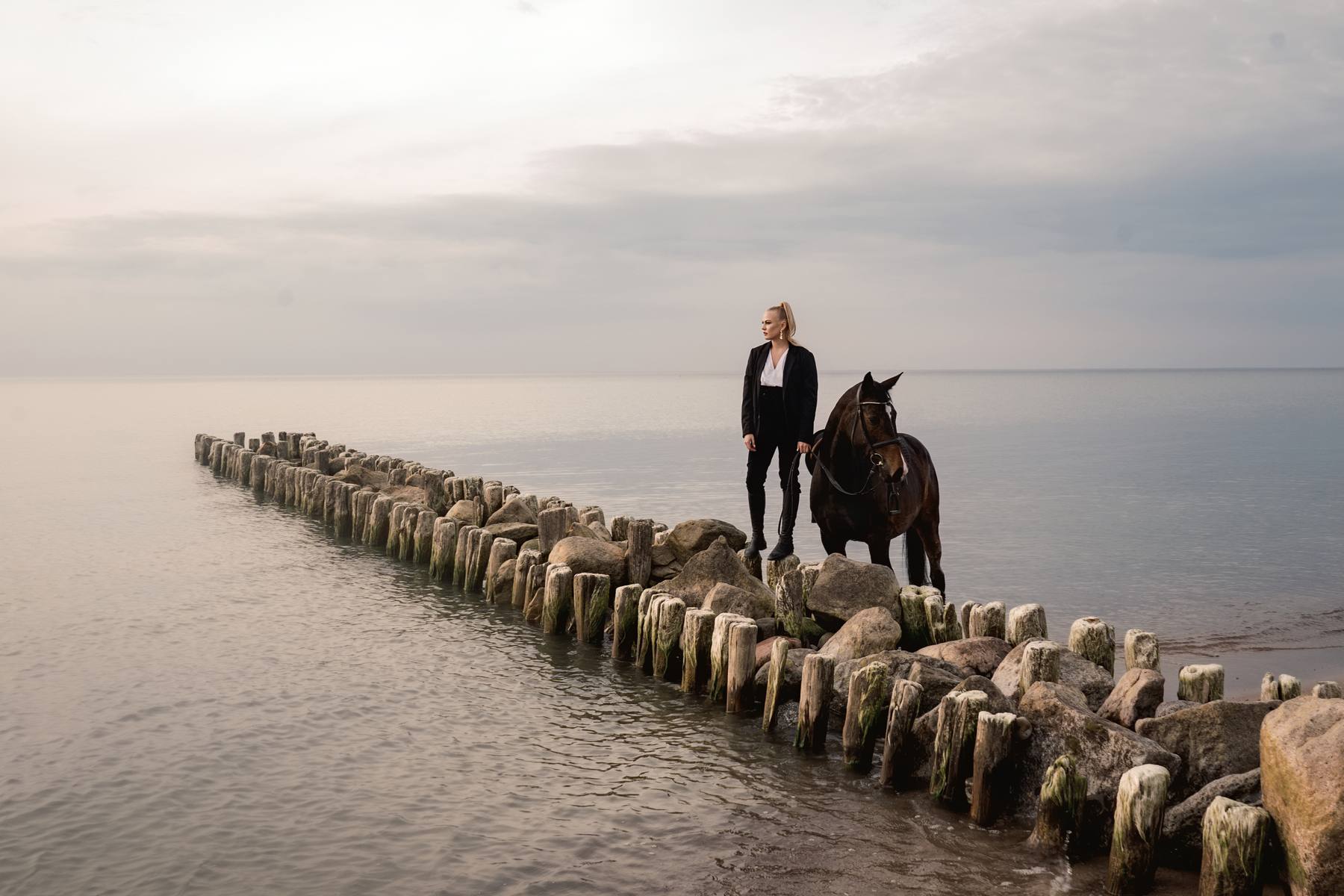 Woman standing with a dark horse at the end of a long timber and stone breakwater, calm gray water and sky