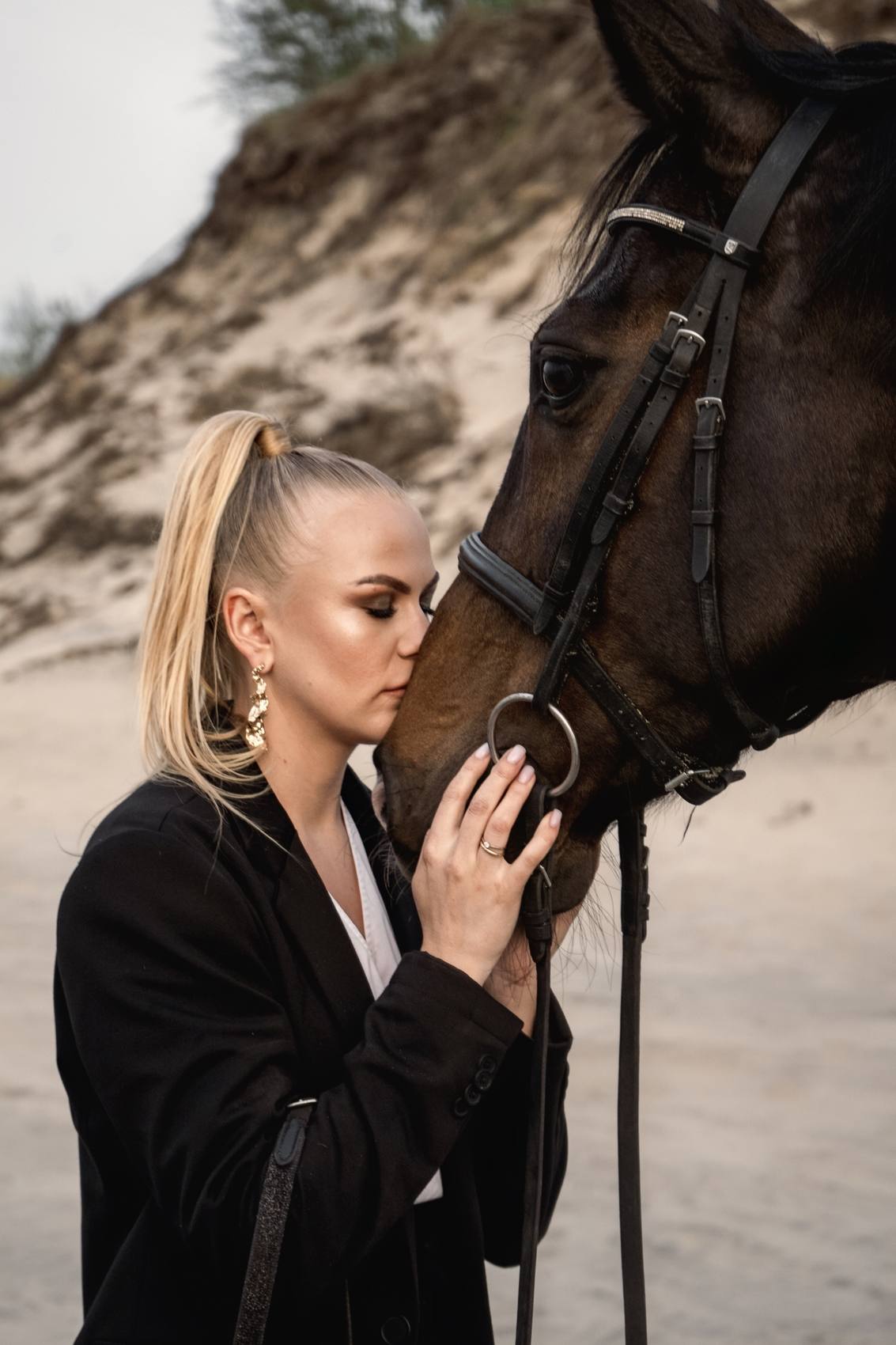 Woman with a blonde ponytail pressing her face to the muzzle of a dark horse, sand dune and grasses behind her