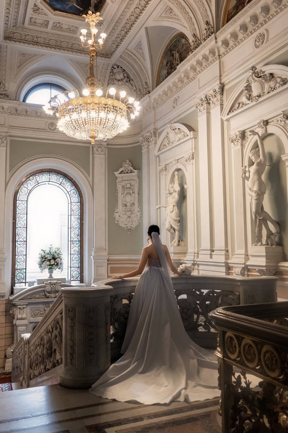 Bride seen from behind with a long veil standing at a curved marble balustrade beneath a crystal chandelier, a stained-glass window and a vase of white flowers beyond