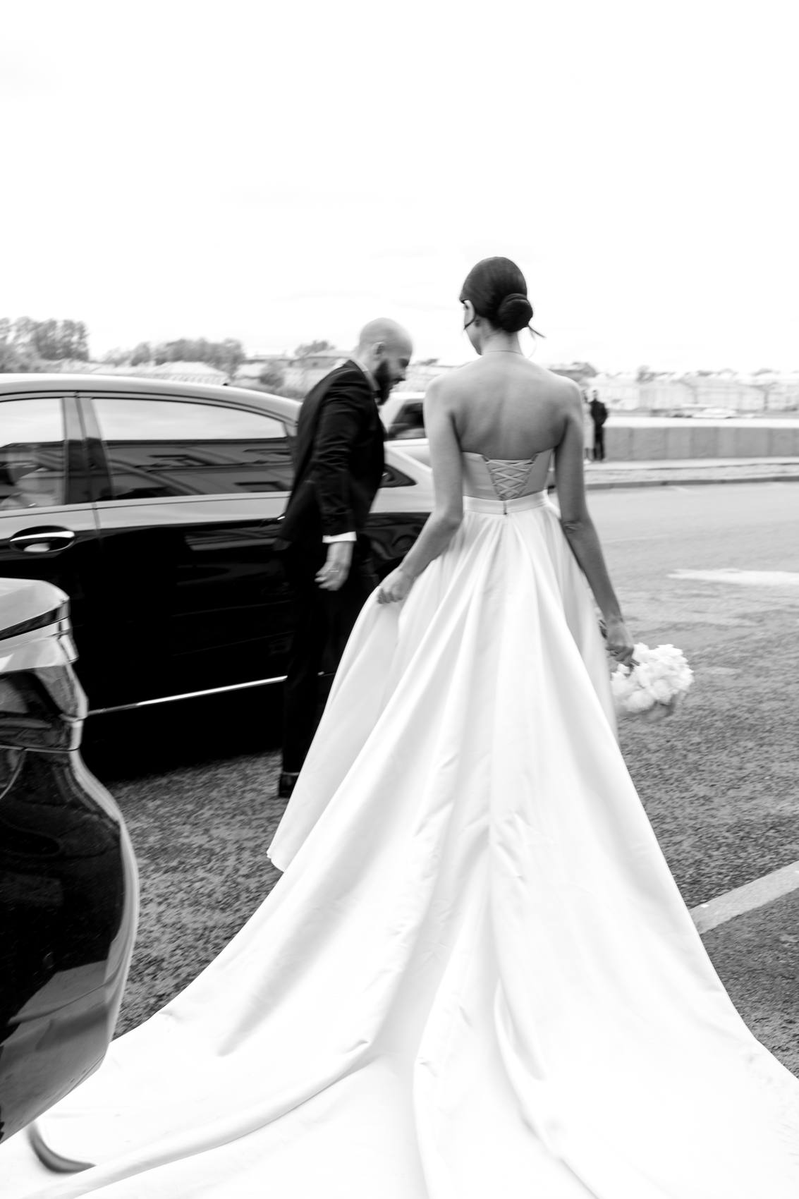 Black and white view from behind of a bride in a corset-back gown with a long train walking toward a black car beside the groom, a bouquet of white flowers in her hand