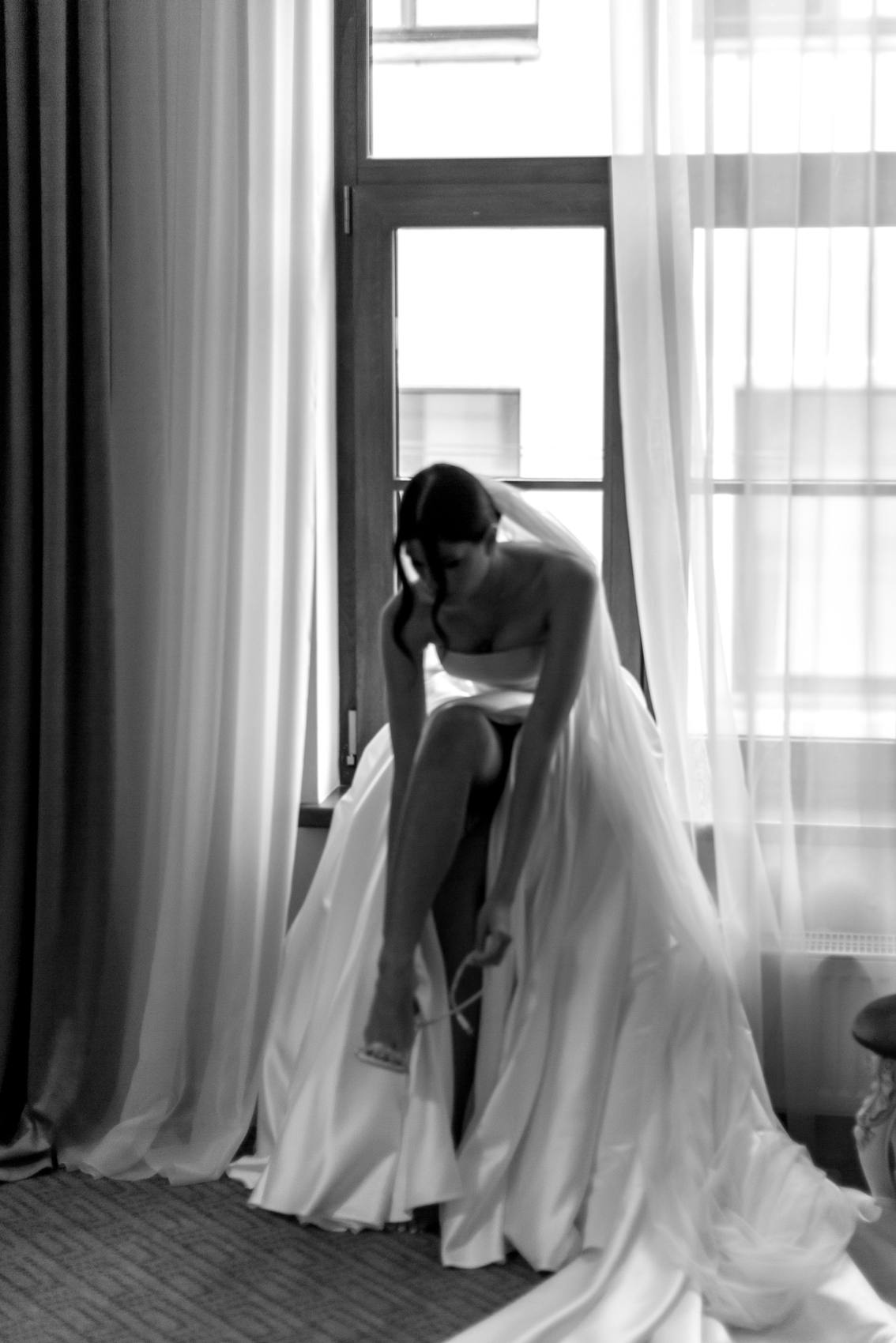 Black and white photograph of a bride sitting on a window sill in a long veil, fastening her sandal beside sheer curtains