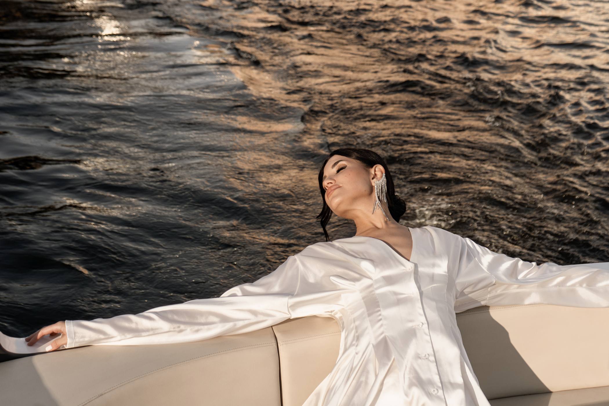 Bride in a white satin dress and long crystal earrings leaning back with her arms spread along the seat of a boat, dark rippling water behind her in warm light