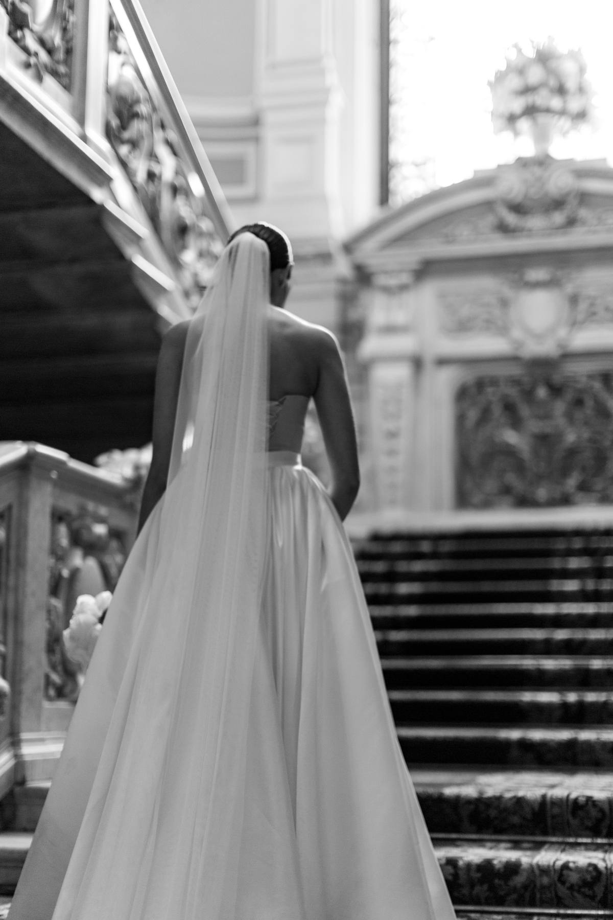 Black and white view from behind of a bride in a long veil and satin skirt facing an ornate staircase with carved stone balustrades