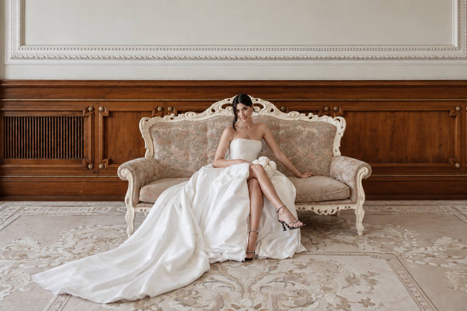 Bride seated on an ivory carved sofa holding white peonies, her legs crossed in jeweled sandals and her long satin train spread across a patterned rug, wood paneling behind her