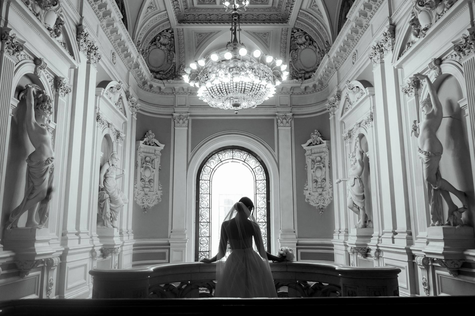 Black and white silhouette of a bride in a long veil leaning on a marble balustrade, facing an arched stained-glass window under a chandelier with statues in niches on both sides