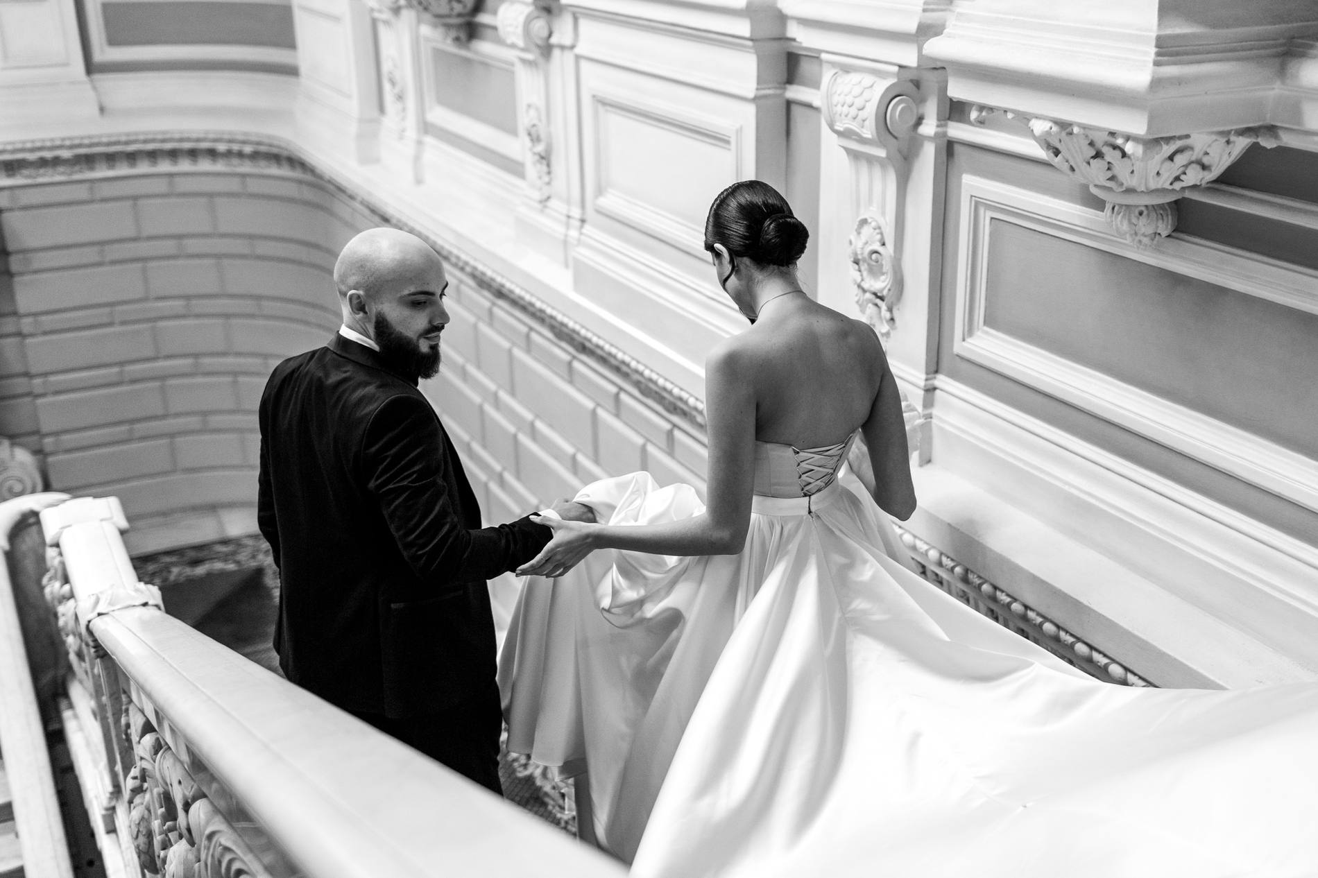 Black and white photograph from above of a groom looking back and holding the bride's hand as she lifts her long satin train on a marble staircase