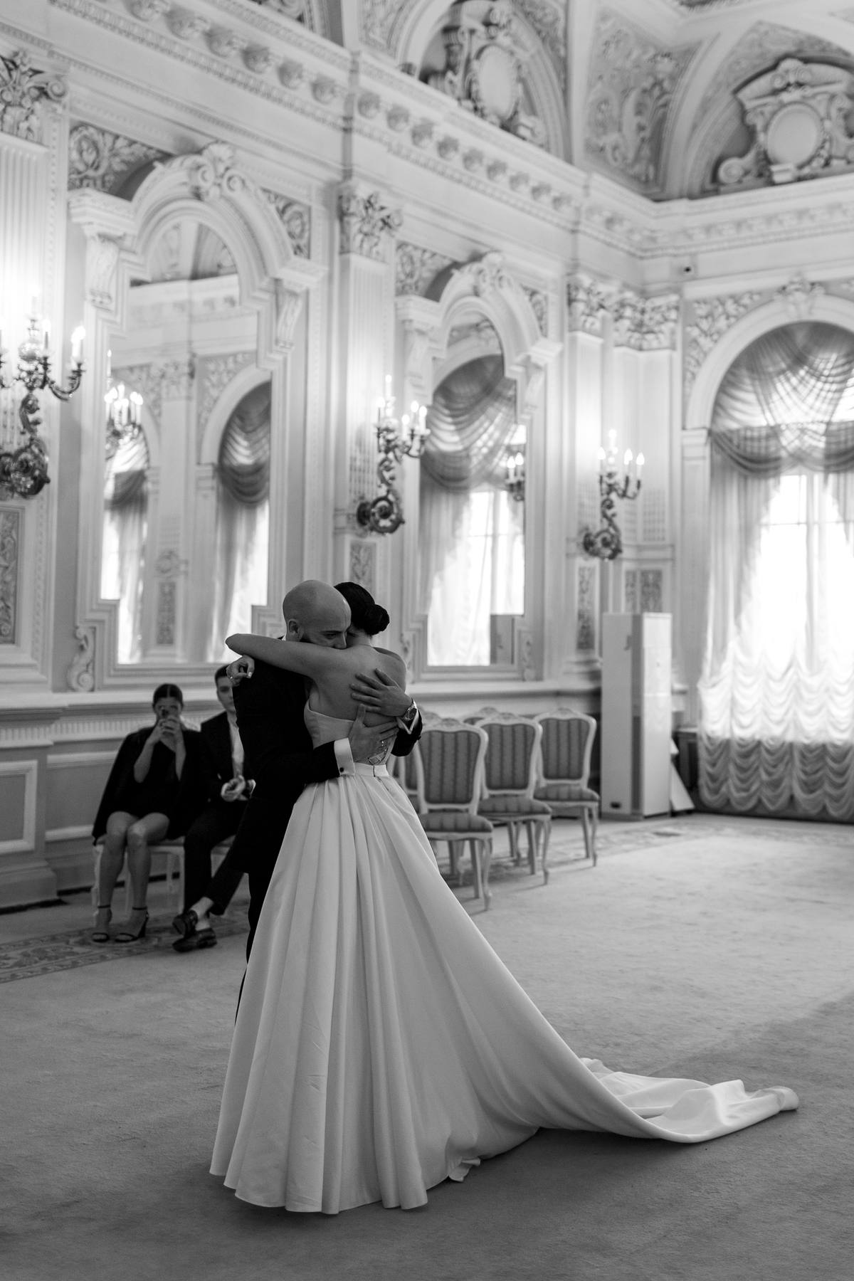 Black and white photograph of a bride and groom embracing in an ornate ballroom with candle sconces and draped windows, two guests seated to the left