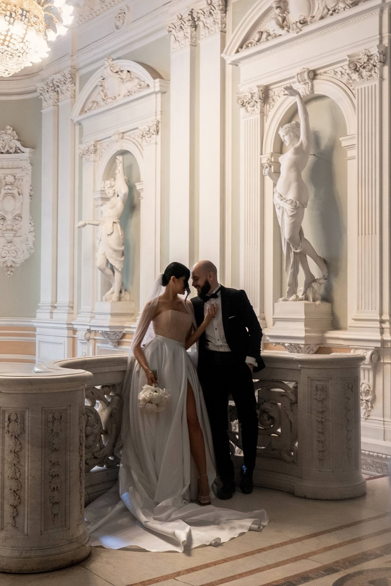 Bride in a strapless gown with a thigh-high slit holding white peonies and groom in a black tuxedo smiling forehead to forehead at a marble balustrade, white statues in arched niches behind them