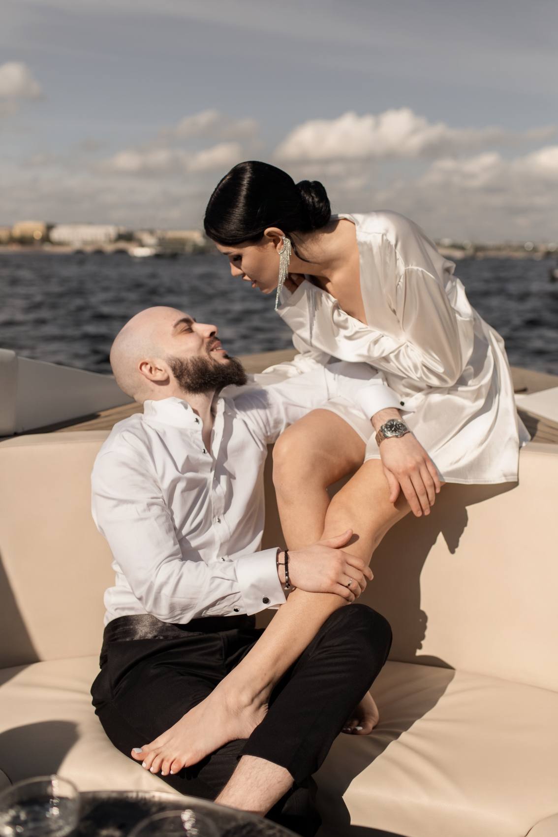 Couple in white seated on a cream boat seat, the bride leaning over the groom as they look at each other, water and a distant shoreline behind under a cloudy blue sky