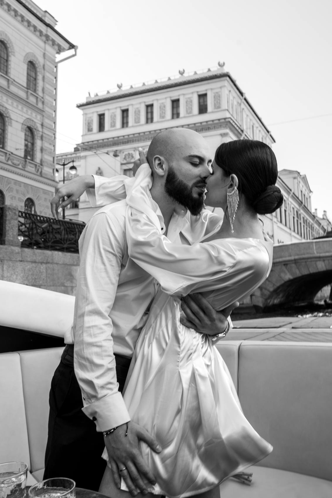 Black and white photograph of a couple kissing on a boat, the bride's arm around his neck, a stone bridge and historic buildings behind them
