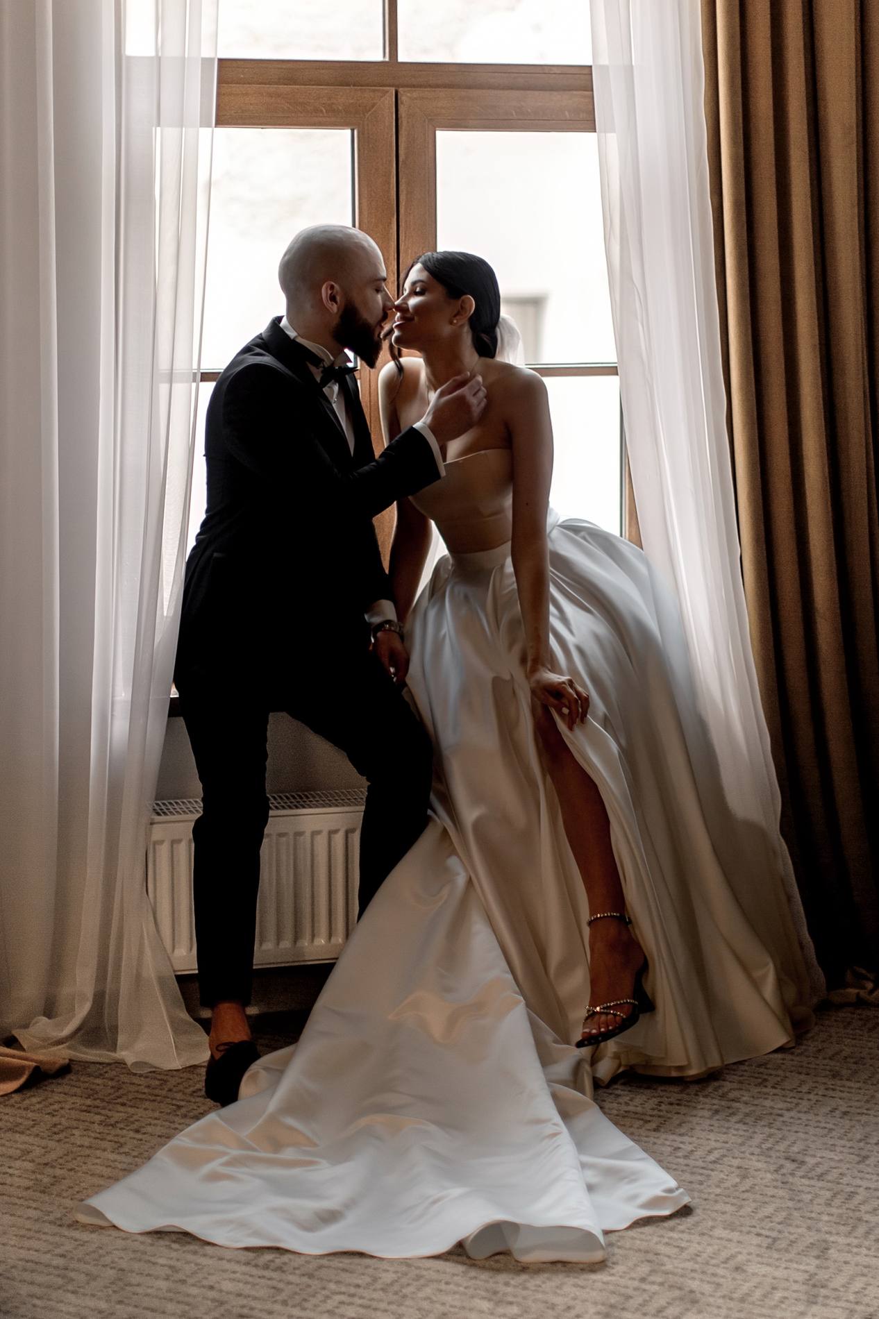Groom in a black tuxedo and bride in a strapless satin gown nose to nose at a tall window with sheer curtains and brown drapes, her train spread across the floor