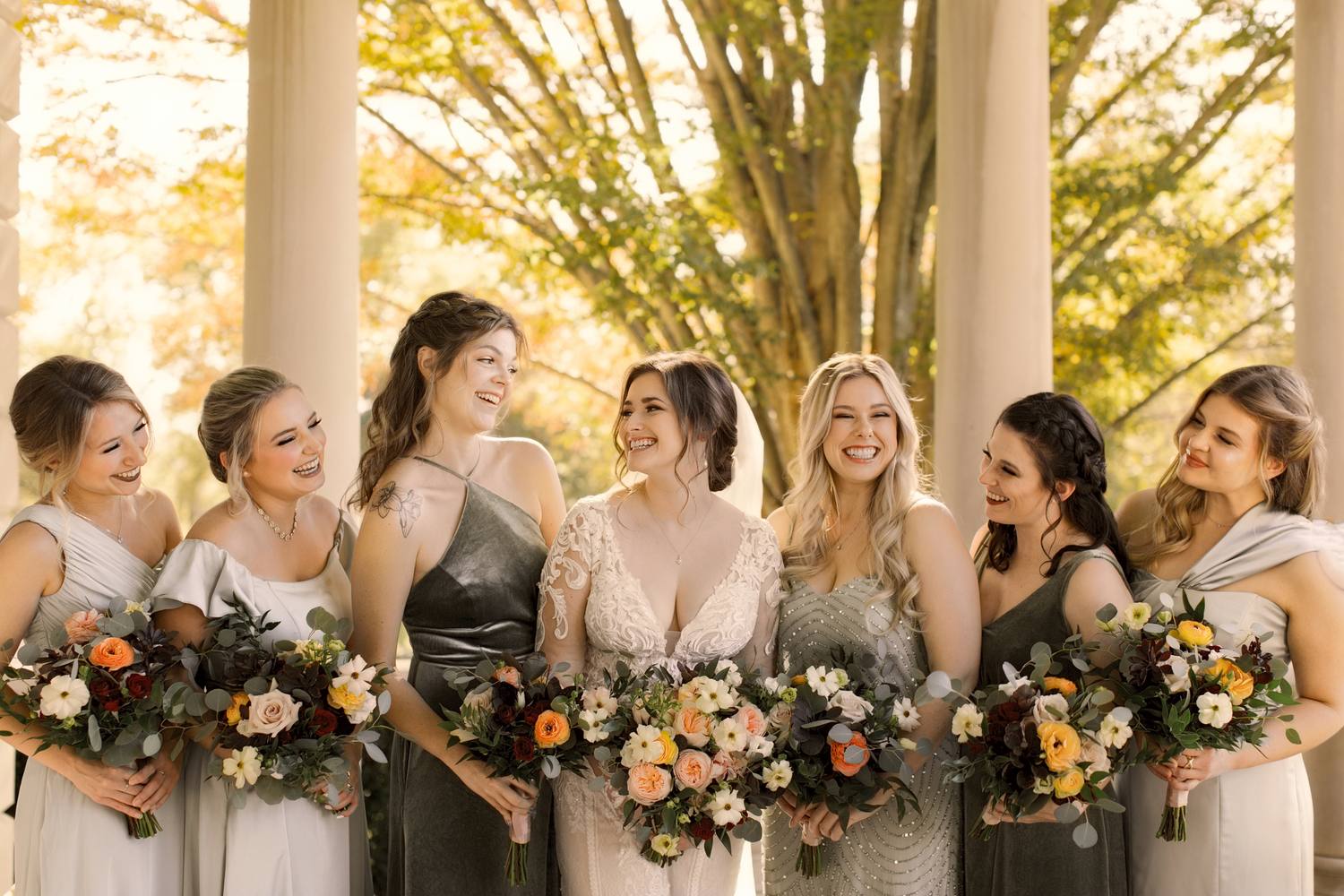 The bride and her bridesmaids laughing and turning toward one another, bouquets in their arms, golden autumn trees behind them