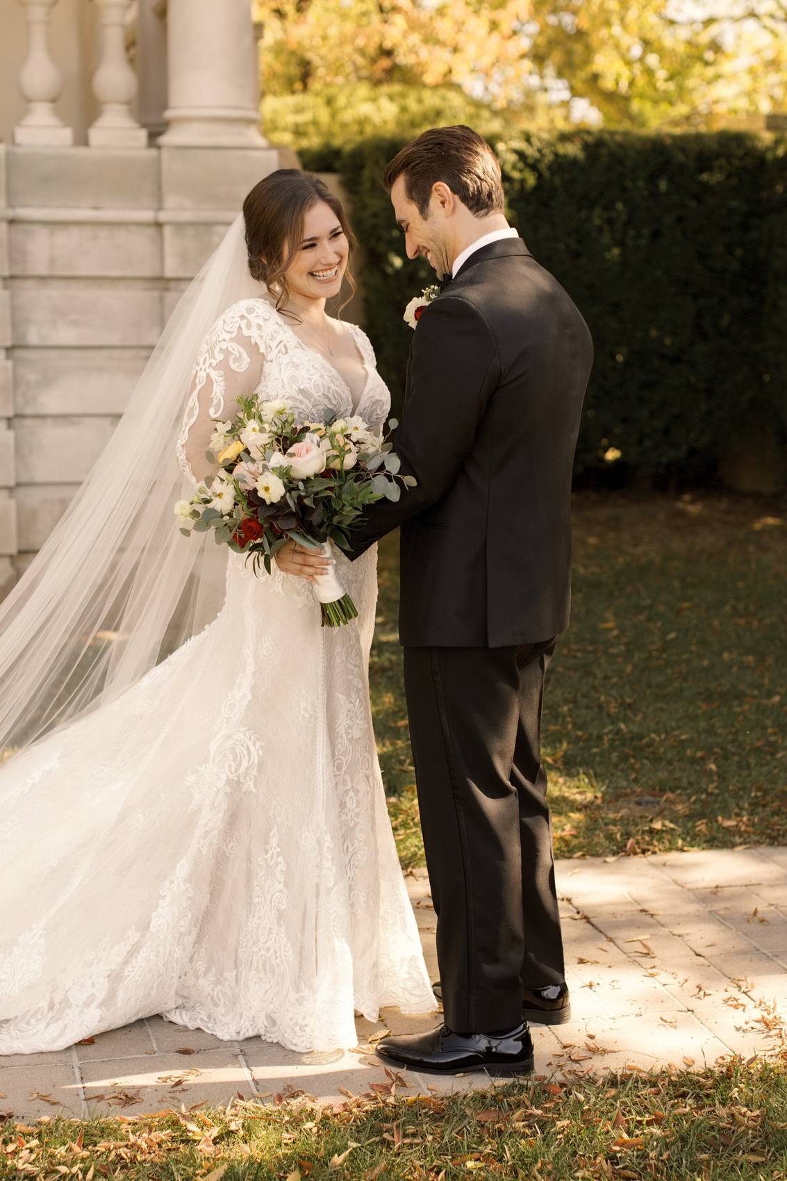 Bride laughing as the groom looks down at her during their first look, her long veil and peach bouquet in the foreground, a clipped hedge and fallen leaves around them
